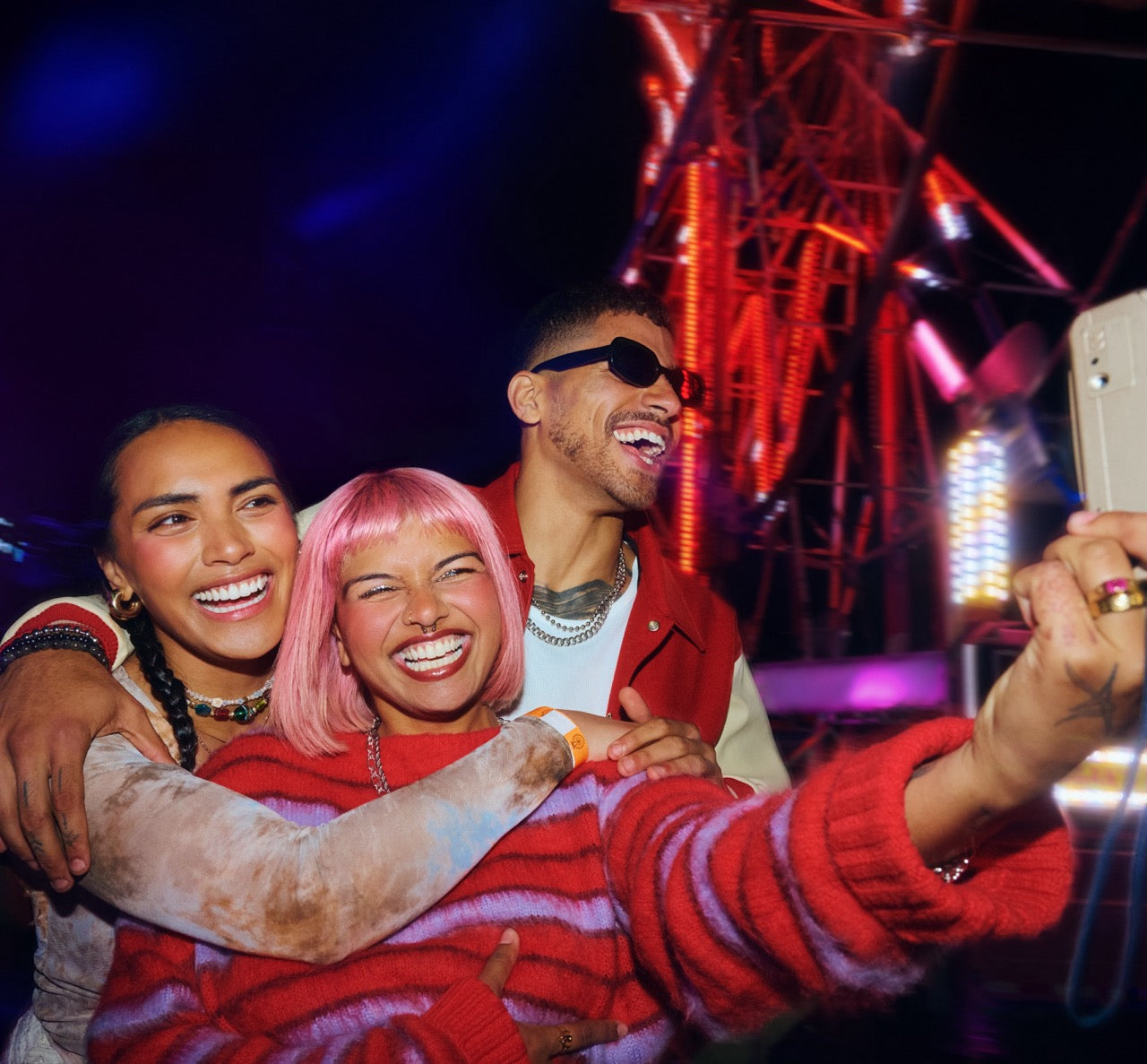 Three friends laughing and taking a selfie at night with colorful lights in the background