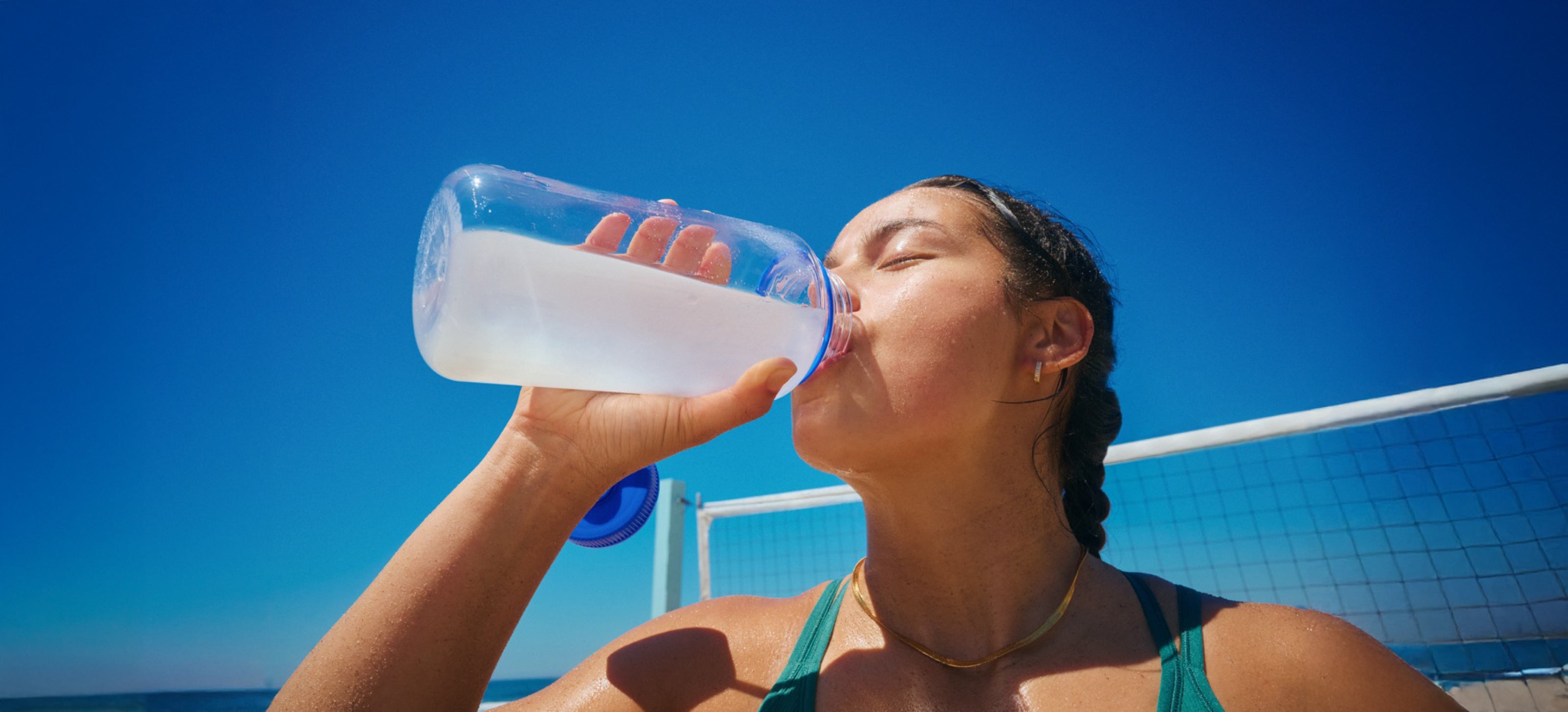 Woman drinking from a water bottle under a bright blue sky, staying hydrated during outdoor activities.